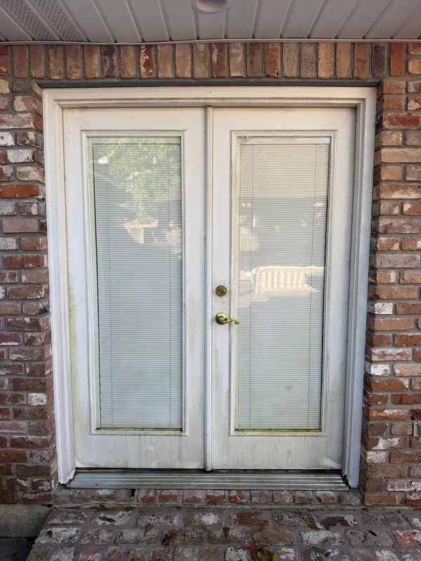 Double white doors with blind covered windows set into a brick wall, with grime gathered along the threshold.