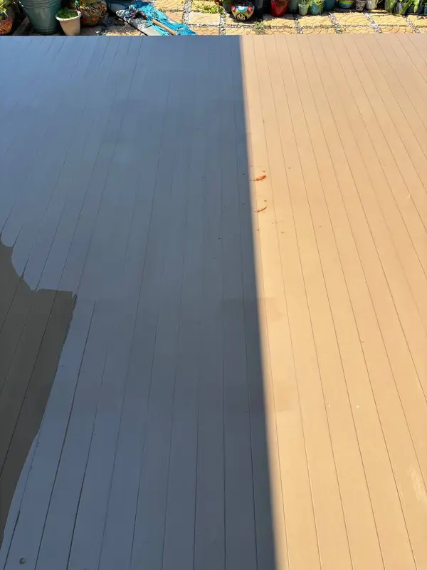 Part of a wooden deck in sunlight next to a deep shadow line, with potted plants and a paved area beyond.