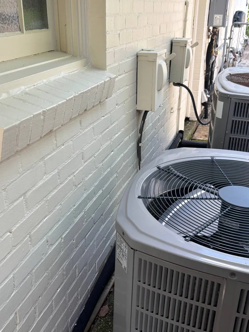 An off white painted brick wall with two electrical disconnect boxes and an air conditioning unit, with green algae between them.