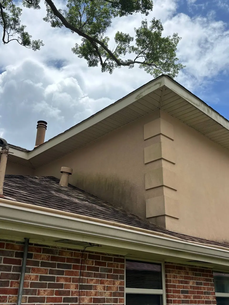 A tan stucco wall with a dark algae streak running down from the eave above a red brick base.