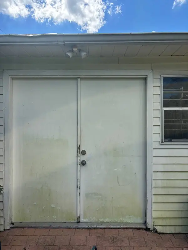 White double doors with green growth along the bottom, a floodlight above and a barred window beside them.