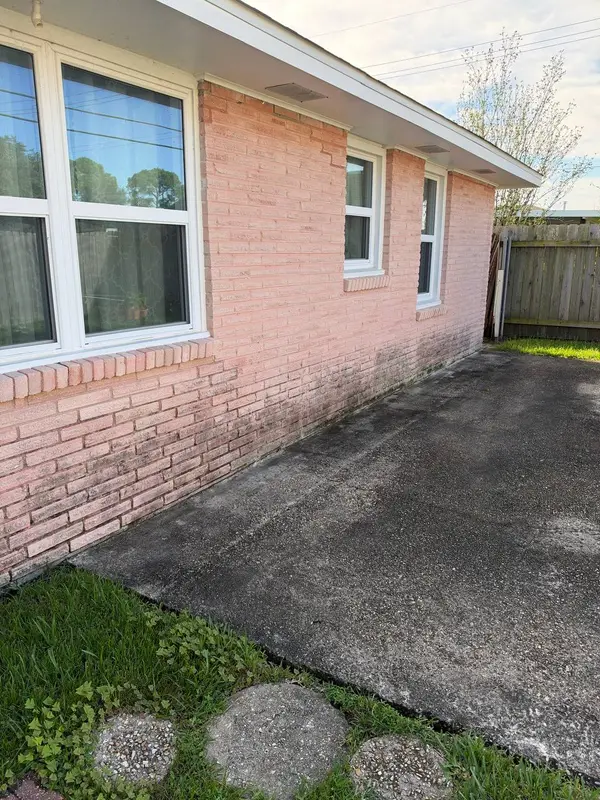 A pink brick wall with two white windows and mildew staining near the base, with a wooden fence beyond.