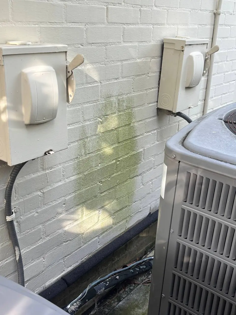 An off white painted brick wall with two disconnect boxes and an air conditioning unit, with algae between the boxes.