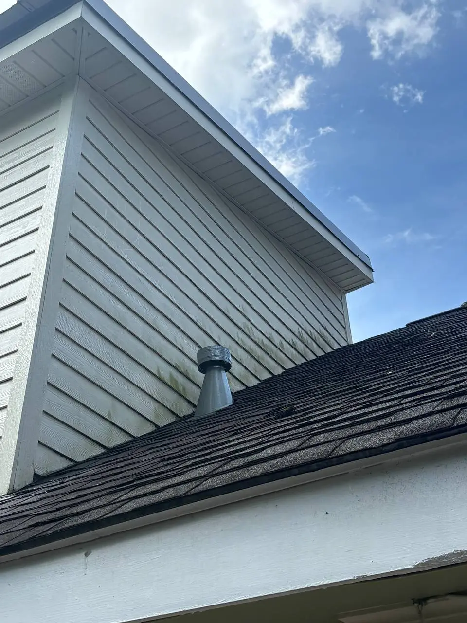 A shingled roof slope in the foreground with a vent pipe, and siding stained with mildew below the roofline.