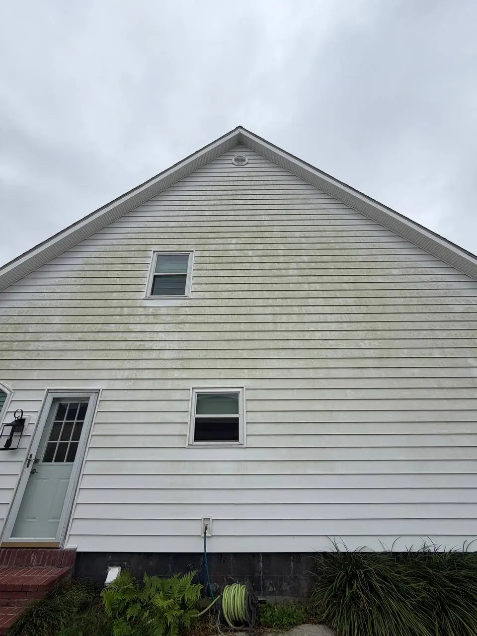 A white sided gable wall with algae streaking, a second storey window and a ground floor door and window under an overcast sky.