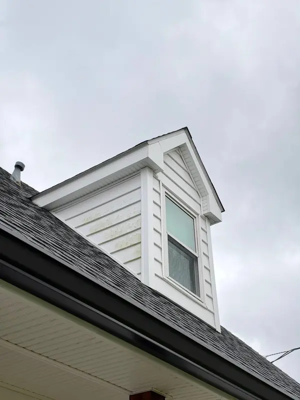 A white sided dormer window set into a dark shingle roof, with algae patches on the siding.