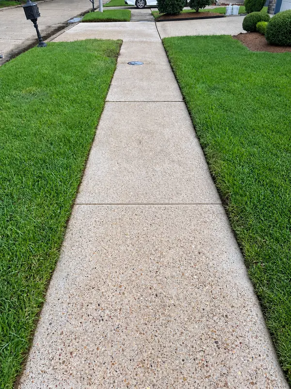 An exposed aggregate concrete sidewalk bordered by grass, with a mailbox and a parked car at the far end.