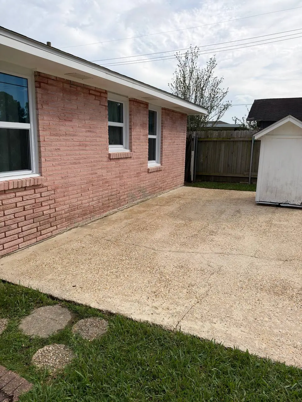 The side of a pink brick building with two windows above a weathered concrete patio and a white shed, under an overcast sky.