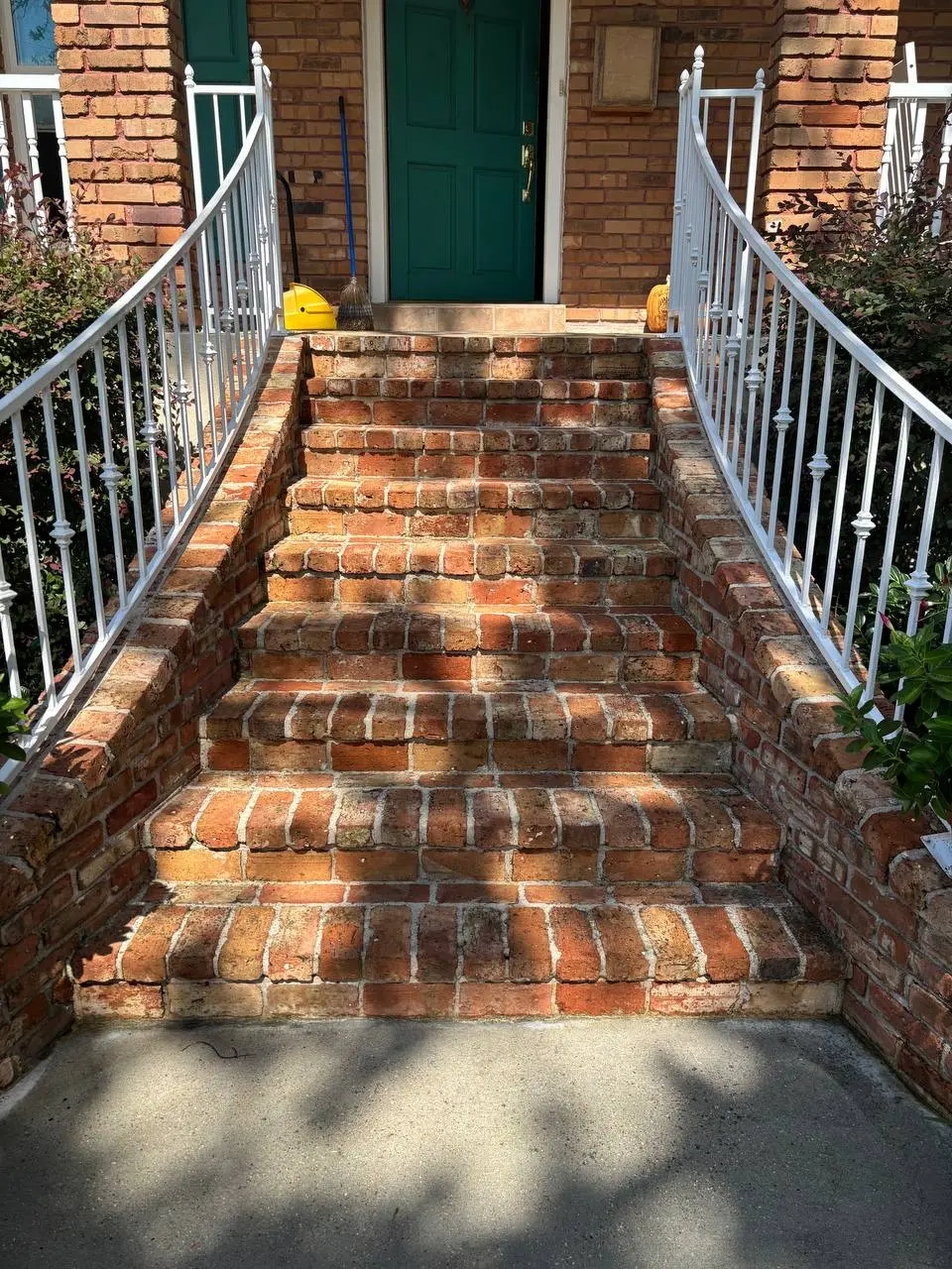 Seven brick steps with white railings leading to a teal door, with a broom and dustpan leaning against the wall.