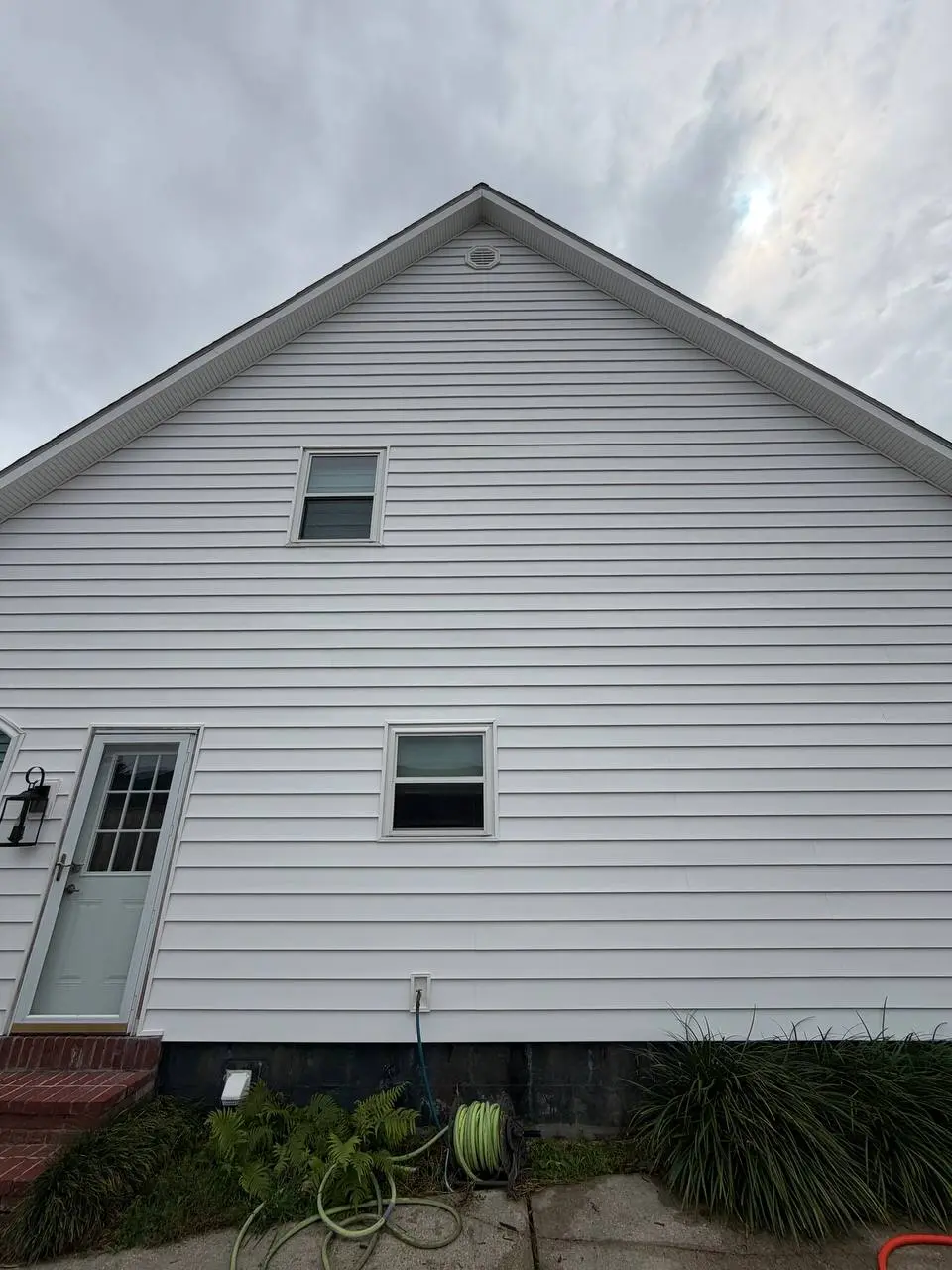 A white sided gable wall with a round vent, two windows and a hose coiled on a reel at the base.