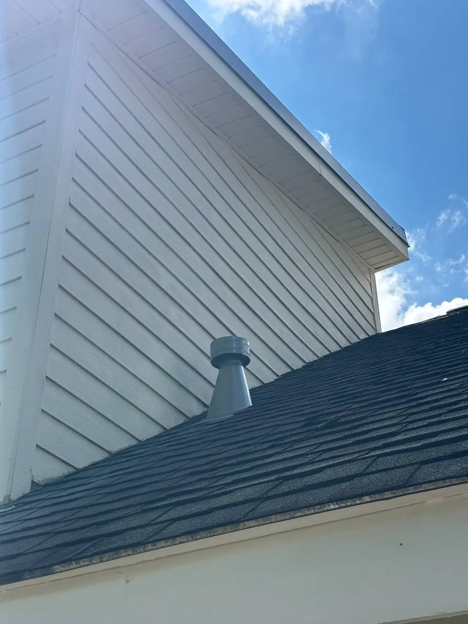 A roof slope with a metal vent stack in the foreground and a white sided gable beyond, against a blue sky.