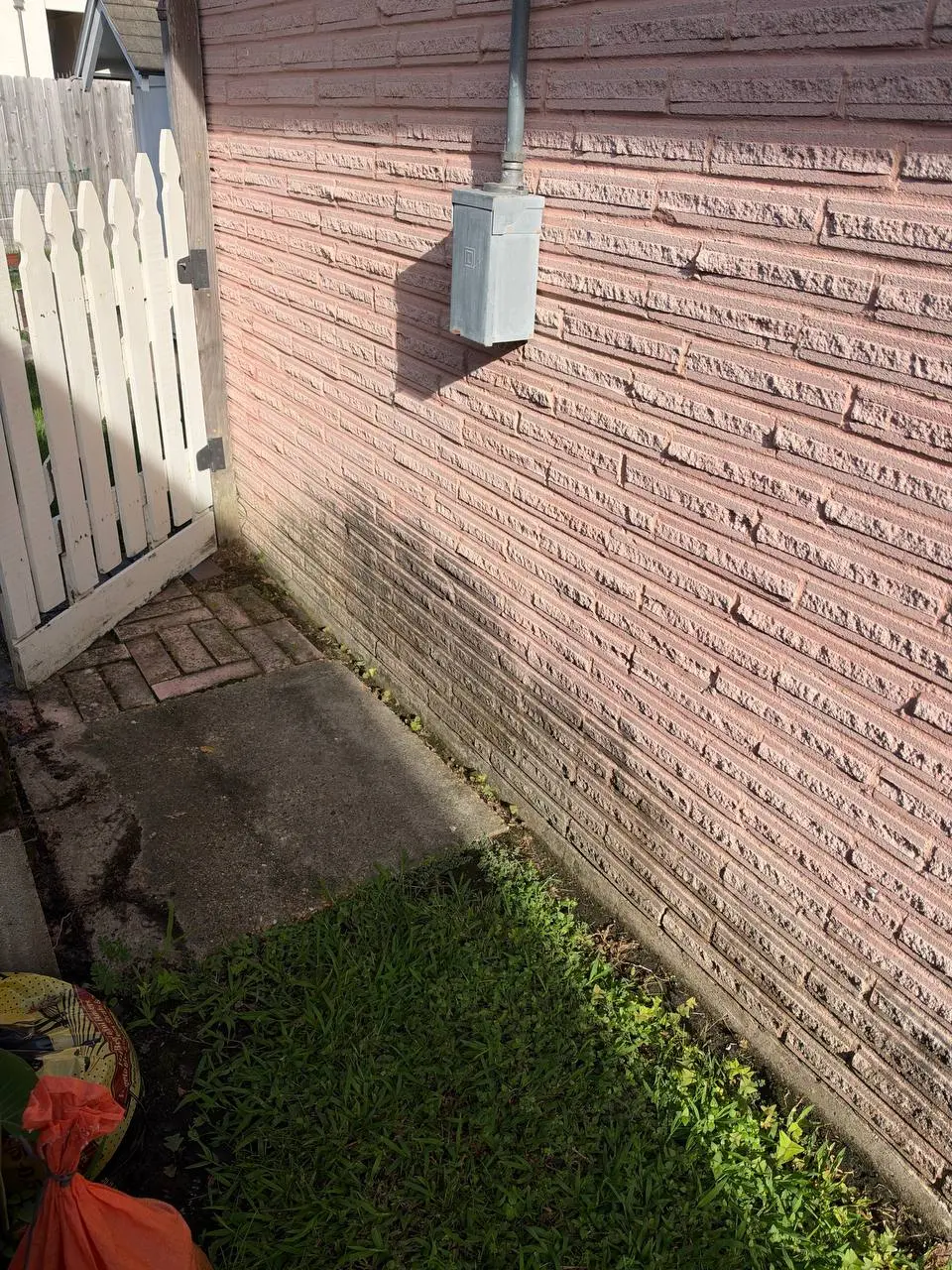 A pale brick wall with grey streaks near the base, a utility box and a white gate beside a concrete slab.