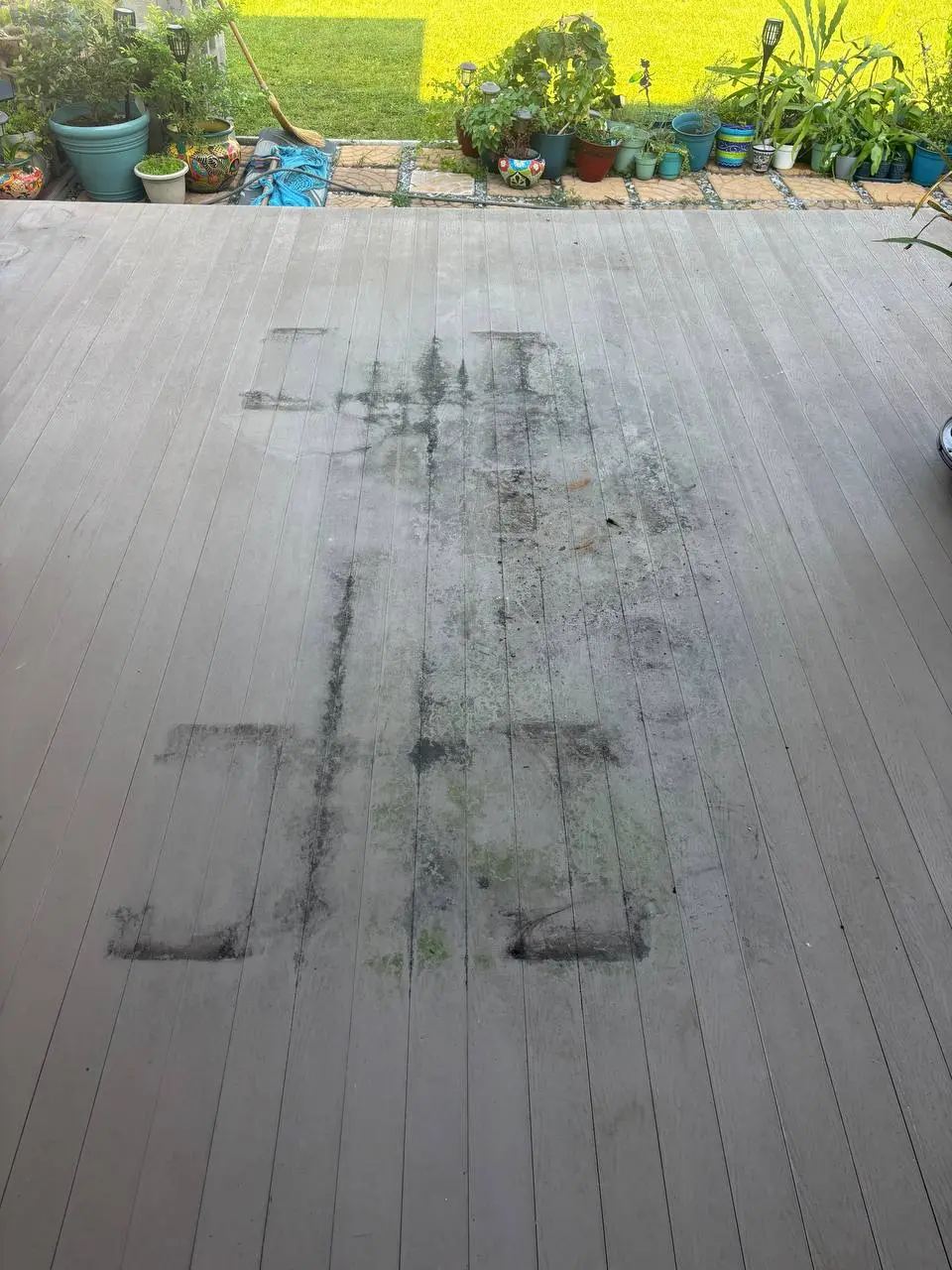 A grey deck marked by dark staining in the shapes where furniture stood, with potted plants beyond.