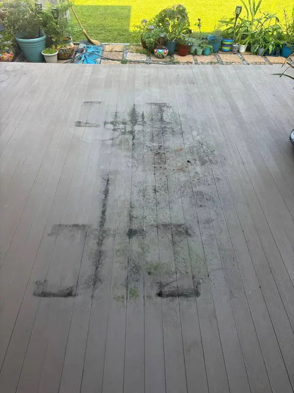 A grey deck marked by dark staining in the shapes where furniture stood, with potted plants beyond.