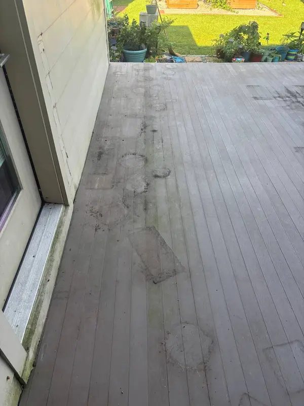 A grey wooden deck with a band of dark staining and clean patches where furniture sat, beside cream siding.