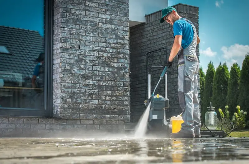 A worker in a cap and overalls using a pressure washer wand on a stone patio, with the machine and a detergent jug beside him.